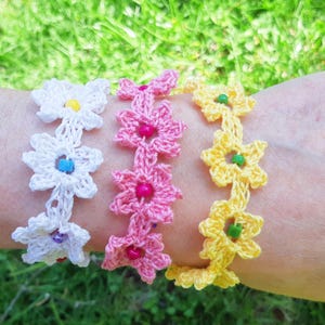 May include: Three crochet flower bracelets in white, pink, and yellow are displayed on a wrist. The white bracelet has blue and purple beads, the pink has pink beads, and the yellow has green beads. The bracelets are handmade.