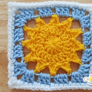 May include: A crocheted square with a yellow sun design in the center. The sun is surrounded by a blue border and a white outer border.