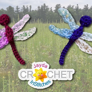 May include: Two crocheted dragonfly ornaments, one in shades of purple and burgundy, the other in shades of blue and white, against a backdrop of a grassy field. The text "Jayda Crochet InStitches" is visible in the lower center of the image.