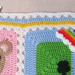 May include: A colorful crochet baby blanket with a bear, rainbow, and green square designs. The blanket has a white, blue, yellow, and white border.