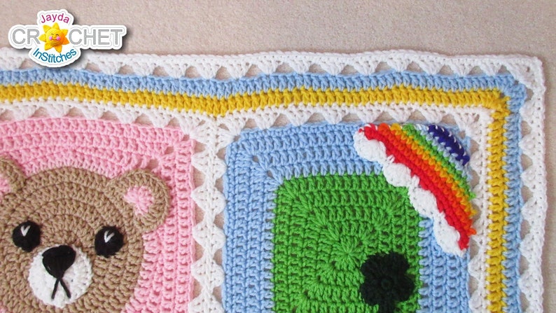 May include: A colorful crochet baby blanket with a bear, rainbow, and green square designs. The blanket has a white, blue, yellow, and white border.