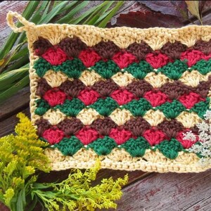 May include: A handmade crochet potholder with a repeating pattern of red, green, and brown diamonds on a beige background. The potholder has a loop for hanging. The image also includes yellow flowers and green grass.