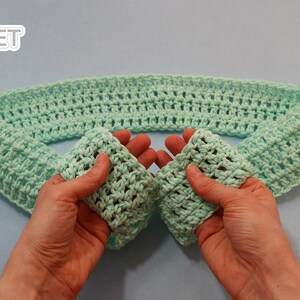 May include: A light green crocheted headband, folded in half, with the ends held in two hands.