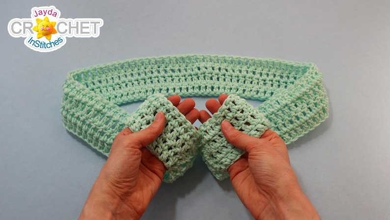May include: A light green crocheted headband, folded in half, with the ends held in two hands.