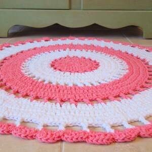 May include: A round, handmade area rug with a crochet design. The rug features concentric circles of white and coral pink. The edge has a scalloped trim. The words "Area Rug" are in a blue bubble font in the upper left corner.