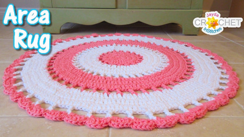 May include: A round, handmade area rug with a crochet design. The rug features concentric circles of white and coral pink. The edge has a scalloped trim. The words "Area Rug" are in a blue bubble font in the upper left corner.