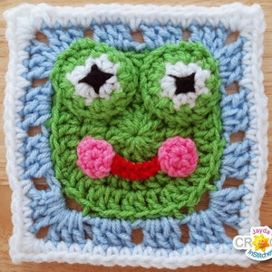 May include: A green crochet square with a frog face. The frog has black eyes, a red mouth, and pink cheeks. The square is bordered with white and light blue crochet.