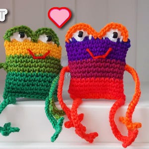 May include: Two handmade crochet frog toys in vibrant colors. One frog is green, yellow, and orange, the other is red, purple, and orange. Both have black eyes and red smiles. A pink heart floats above them. The logo "Jayda Crochet InStitches" is in the top left.