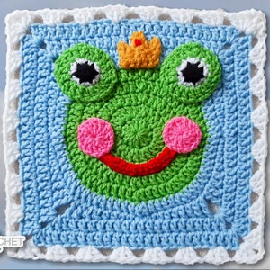 May include: A crocheted square featuring a green frog with a yellow crown, pink cheeks, and a red smile. The frog is surrounded by a blue crocheted border with a white crocheted edge.