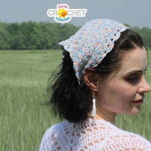 May include: A woman wearing a crocheted headband in shades of blue, pink, and white. The headband is tied in a knot at the back of her head. She is standing in a field of green grass.
