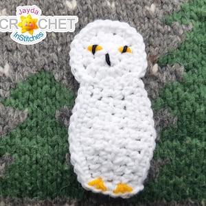 May include: White crocheted owl with yellow eyes and feet. The owl is sitting on a gray and green crocheted background.