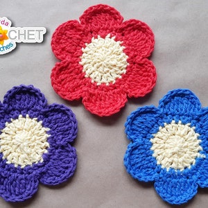 May include: Three crocheted flower coasters in red, purple, and blue with a cream colored center. The coasters are on a brown surface with green vines.