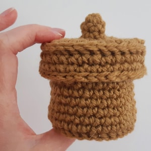 May include: A crocheted acorn-shaped container with a brown lid and a brown body. The container is small enough to fit in the palm of a hand.