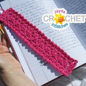 Op de afbeelding: Een roze gehaakte bladwijzer met een kwast. De bladwijzer ligt op een notitieboek met handschrift. De tekst op het notitieboek luidt "Jayda CROCHET InStitches".