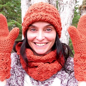 May include: A woman wearing a crocheted hat, scarf, and mittens in a burnt orange color. The hat and scarf are a simple stitch pattern, while the mittens have a more intricate design.