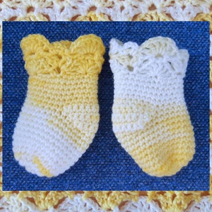 Könnte beinhalten: Ein Paar gehäkelte Babyschuhe in Gelb und Weiß mit einem gewellten Rand. Die Schuhe liegen auf einem blauen Hintergrund.