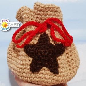 May include: A crocheted brown bag with a red drawstring and a brown star on the front. The bag is tied with a bow. The bag is made by Jayda Crochet in Stitches.
