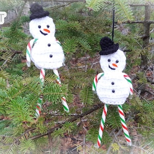 May include: Two white crocheted snowman ornaments hanging from a pine tree branch. Each snowman has a black top hat, black eyes, an orange carrot nose, and a red and white candy cane for arms. The candy canes are green and red striped.