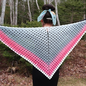 May include: A handmade crochet shawl in a triangular shape, featuring a gradient of colors. The shawl transitions from a white border to a pink section, then to a gray center. The edges have a scalloped design.