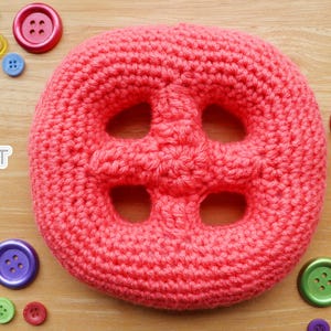May include: A large, round, coral-colored crochet button with four central holes, surrounded by various small, colorful plastic buttons. The text "Jayda InStitches" with a sun graphic is visible on the left side of the image.