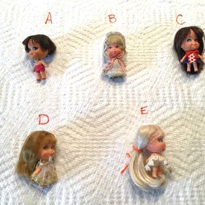 May include: Five small vintage dolls with various hairstyles and outfits are arranged on a white textured surface. The dolls have painted faces and are wearing dresses or outfits in different colors. The dolls are in good condition and appear to be collectibles.
