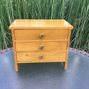 May include: A small wooden dresser with four drawers and brass knobs. The dresser is light brown and has a simple design.