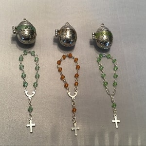 May include: Three silver egg-shaped charms with the words "Joy", "Blessed", and "Peace" on them. Each charm is attached to a small silver chain with a cross at the end. The chains have green, amber, and green beads.
