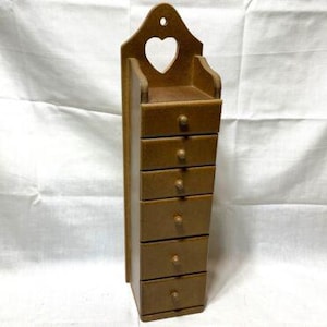 Peut inclure: Un rangement mural en bois avec une découpe en forme de cœur en haut. L'unité a six tiroirs, chacun avec une simple poignée.