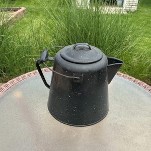 May include: A black speckled enamelware coffee pot with a lid, spout, and handle. The pot is sitting on a round table with a woven edge, with green grass in the background.