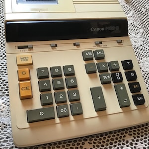 May include: A vintage Canon P115-D calculator with a beige body and black keys. The calculator features a numeric keypad, function keys, and a paper feed button. The brand name is visible on the top.