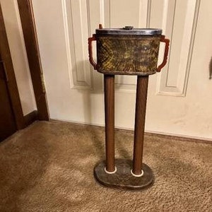 May include: A vintage, wooden, freestanding ashtray with a green and brown marbled top and two red plastic handles. The ashtray has a silver lid and stands on two fluted columns.