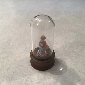 May include: A small figurine of a woman in a blue and pink dress holding an apple is displayed under a glass dome on a wooden base.