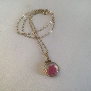 May include: A gold chain necklace with a round locket pendant. The locket is open and contains small, pink, square-shaped pieces of glass.