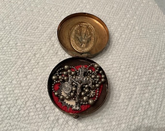 May include: An open, antique brass-coloured round box. The lid features a religious medallion. Inside, a rosary with a crucifix and a small religious medal rests on a red fabric lining.