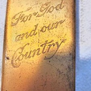 May include: A rectangular, weathered gold-toned metal tin featuring the engraved text "For God and our Country" in a cursive script. The surface shows signs of age, including scratches, scuffs, and a mottled patina across the metallic finish.