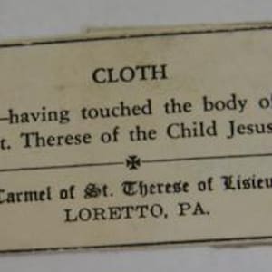 May include: A small, rectangular card with the word "CLOTH" at the top. The text below reads "-having touched the body of St. Therese of the Child Jesus." The card also includes the text "Carmel of St. Therese of Lisieux LORETTO, PA."