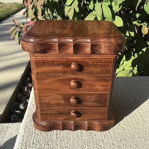 May include: A small, antique wooden jewelry box with four drawers and a decorative top. The box is made of dark brown wood and has round knobs on each drawer. The box is sitting on a concrete surface.