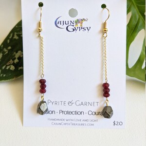 May include: A pair of gold dangle earrings with a pyrite and garnet stone at the bottom. The earrings are on a white card with the text "PYRITE & GARNET", "sion - Protection - Coura", "HANDMADE WITH LOVE AND LIGHT", "CAJUNGYPSY TREASURES.COM", and "$20".