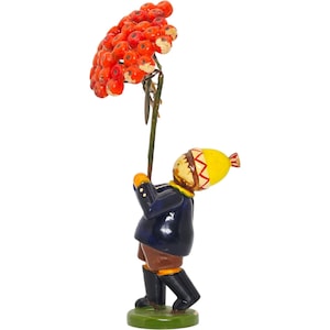 Könnte beinhalten: Eine skurrile Figur einer Person, die eine Traube leuchtend orangefarbener Beeren hält. Die Figur trägt einen marineblauen Mantel, eine braune Hose und schwarze Stiefel mit einer gelben Mütze. Der Sockel ist grün.