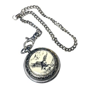 May include: Silver pocket watch with a chain. The watch face has a white background with a black and white image of an eagle in flight.