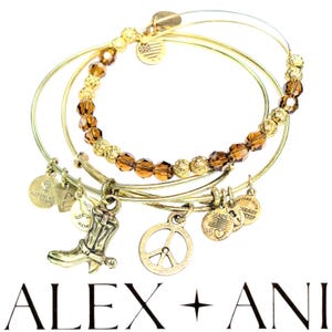 May include: Set of three gold bangle bracelets with brown and gold beads. The bracelets have charms including a peace sign, a boot, and a heart with the words "Made with Love" and "Infused with Energy" on it. The bracelets are from the brand Alex and Ani.