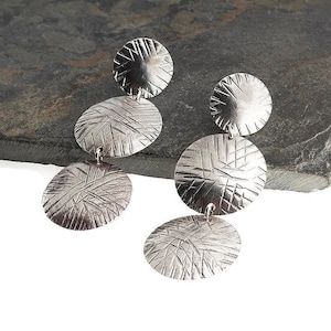 May include: Silver-toned dangle earrings with a textured, circular design. Each earring features three graduated circles, connected by small loops. The surface of each circle has a crisscross pattern, creating a unique visual texture. The earrings are displayed against a dark gray background.