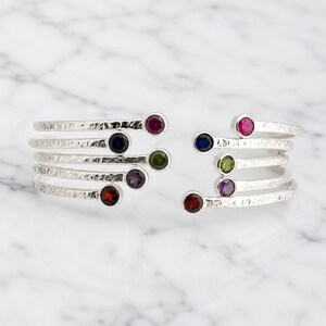 May include: Two silver-toned cuff bracelets with a textured finish. Each bracelet features five colorful gemstones: red, blue, green, purple, and a second red. The bracelets are displayed on a white marble surface.