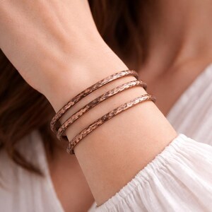 May include: Three copper-colored bangles with a hammered texture are stacked on a wrist. The bracelets have a rustic, handcrafted appearance. The person is wearing a white long-sleeved shirt.