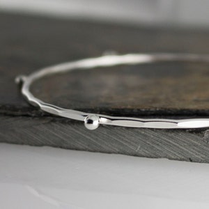 May include: A silver bangle bracelet with a hammered texture and a small, round bead. The bracelet is made of shiny metal and is displayed on a dark gray surface. The bangle is a simple, elegant piece of jewelry.