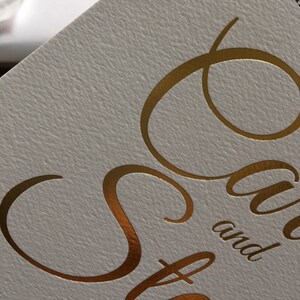 Op de afbeelding: Gouden folielettering op een witte kaart met de tekst "Save the Date" en de namen "Sara en Steve" eronder.