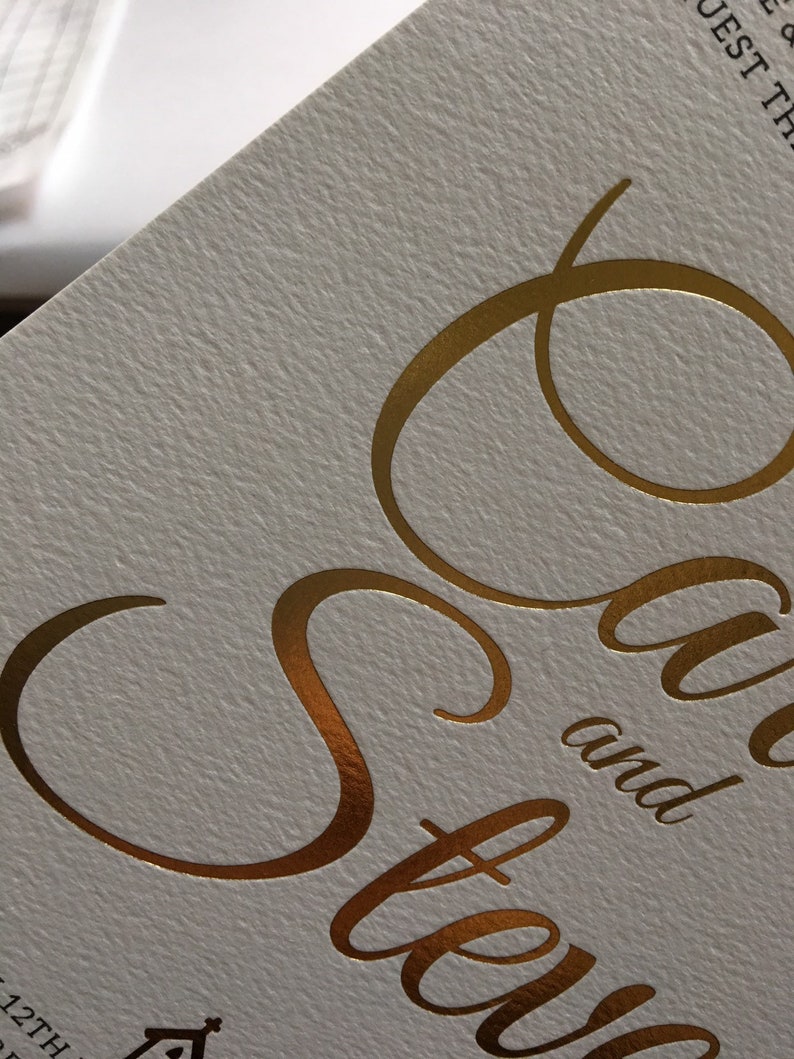 Op de afbeelding: Gouden folielettering op een witte kaart met de tekst "Save the Date" en de namen "Sara en Steve" eronder.