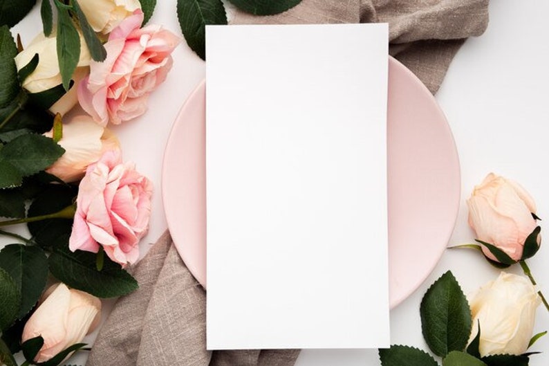 May include: A blank white card on a pink plate with a brown linen napkin and pink roses around it.