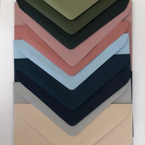May include: A stack of eight envelopes in various colours, including green, dark green, pink, light blue, dark blue, grey, and beige. The envelopes are arranged in a graduated pattern, with the largest envelope at the bottom and the smallest at the top.