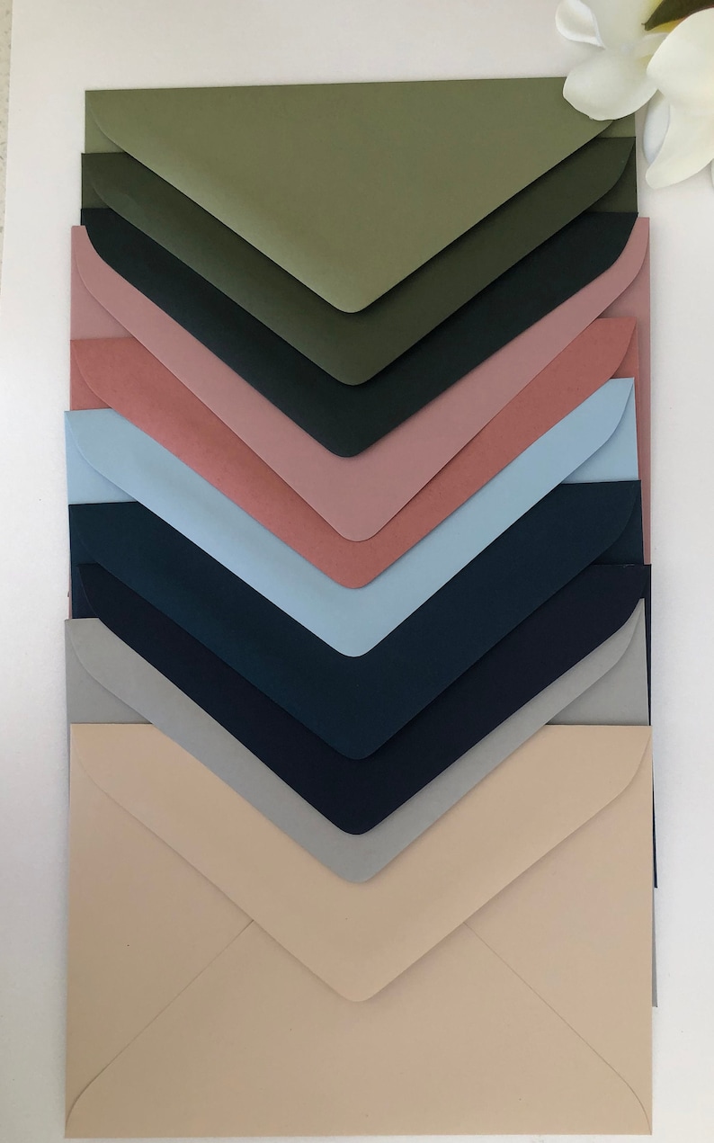 May include: A stack of eight envelopes in various colours, including green, dark green, pink, light blue, dark blue, grey, and beige. The envelopes are arranged in a graduated pattern, with the largest envelope at the bottom and the smallest at the top.